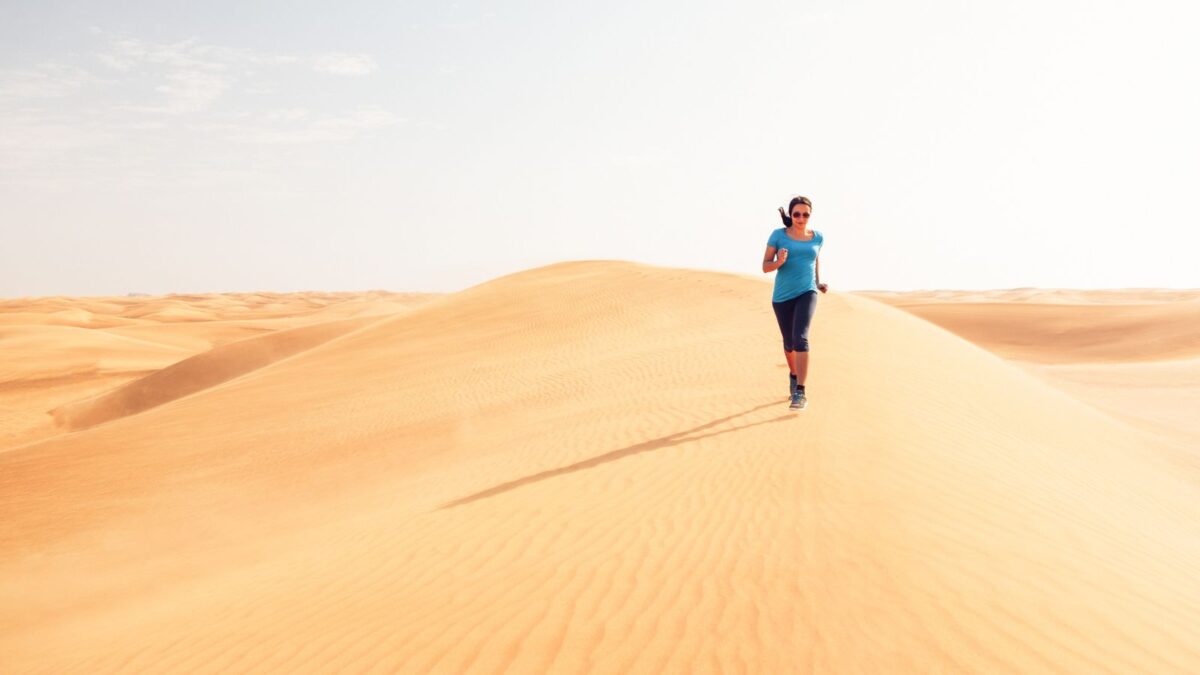 A runner in blue is jogging along the crest of a golden sand dune under a clear sky, with the vast expanse of the desert stretching to the horizon, illustrating isolation and the vastness of nature.