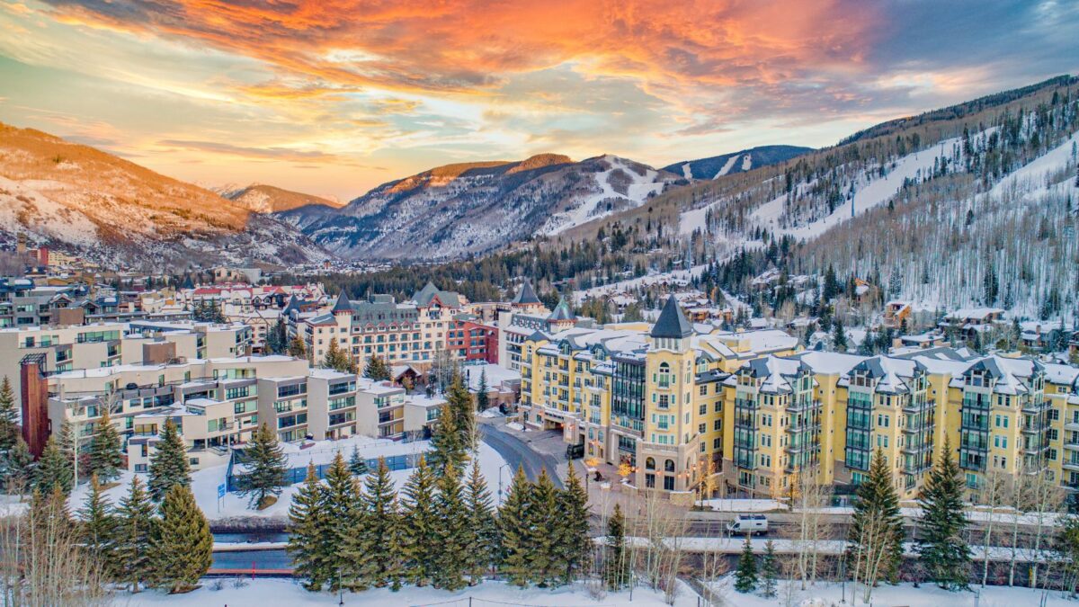 Snow-covered mountain town with colorful buildings at sunset, surrounded by pine trees and snowy peaks.