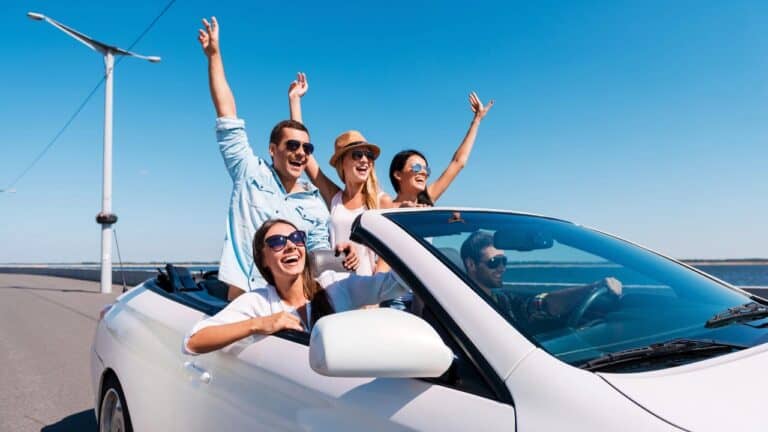 Group of friends enjoying a ride in a white convertible on a sunny day, raising their arms and smiling.