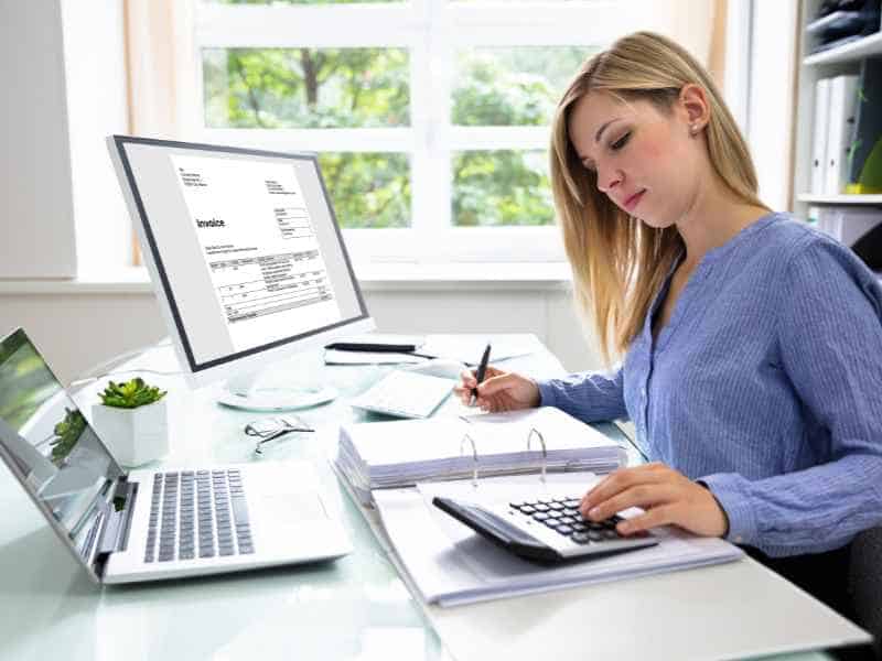 A focused professional working at her desk with a calculator, reviewing financial documents on a computer screen.