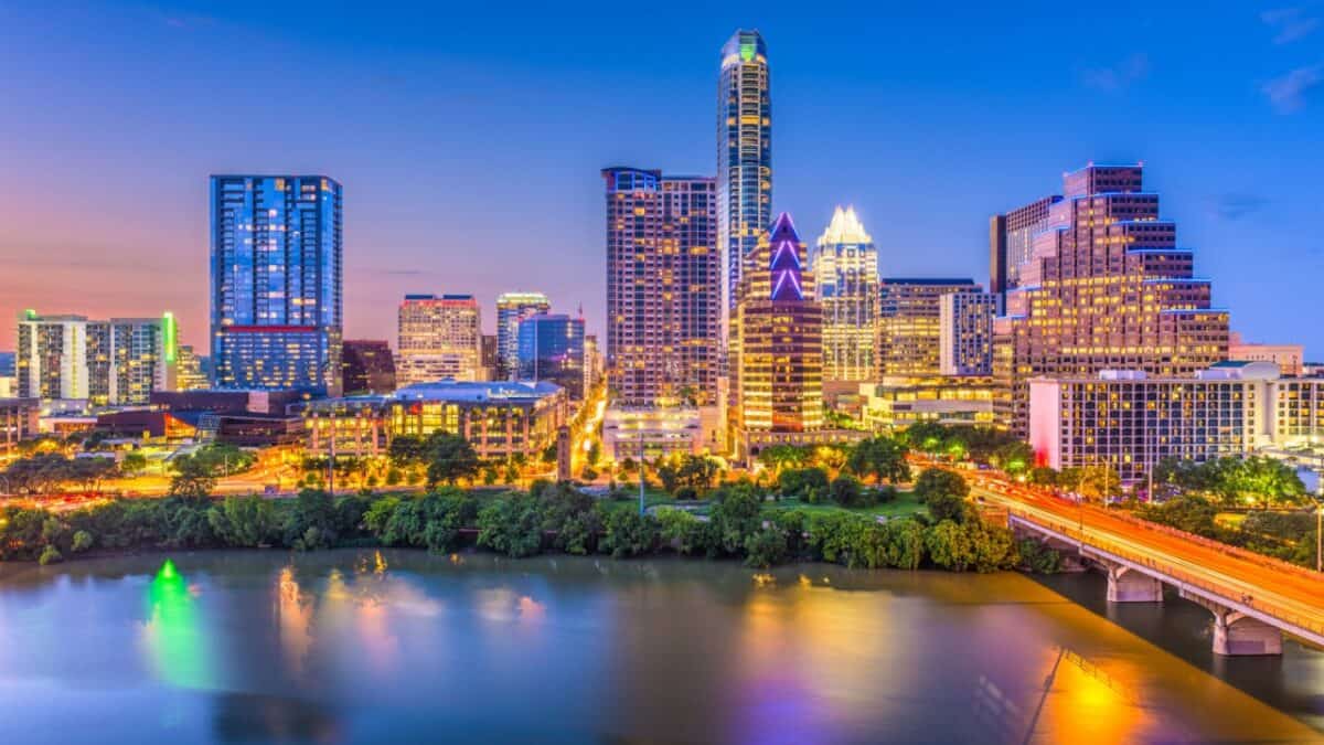 A vibrant sunset skyline: modern skyscrapers towering over a tranquil, reflective river with a bridge leading into the illuminated heart of a bustling city.