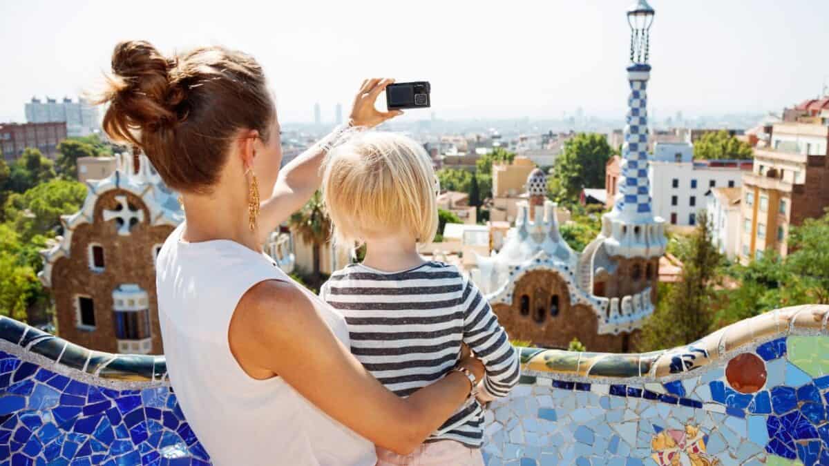 A woman holding a child takes a selfie with a smartphone overlooking the colorful mosaic architecture of Park G&uuml;ell in Barcelona, Spain.