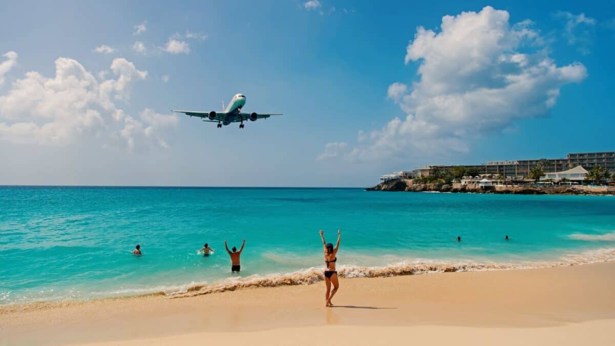 An airplane landing closely over a tropical beach where people are swimming and a woman on the shore is raising her arms in excitement, with clear turquoise water and a blue sky with clouds in the background.