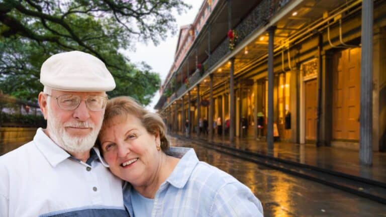 Senior couple smiling and embracing, with a background of an old-fashioned street lined with historical buildings, possibly suggesting a nostalgic trip or a leisurely stroll through a charming district.