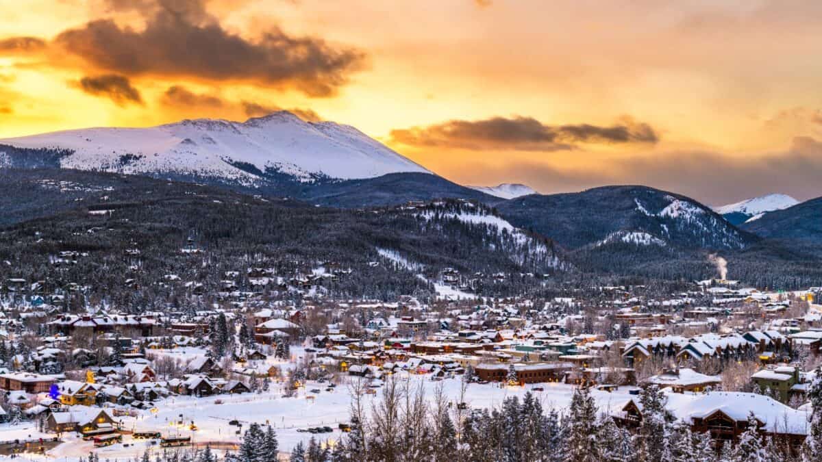 Snowy mountain village at sunset with orange and yellow sky, surrounded by forested hills and snow-covered rooftops.