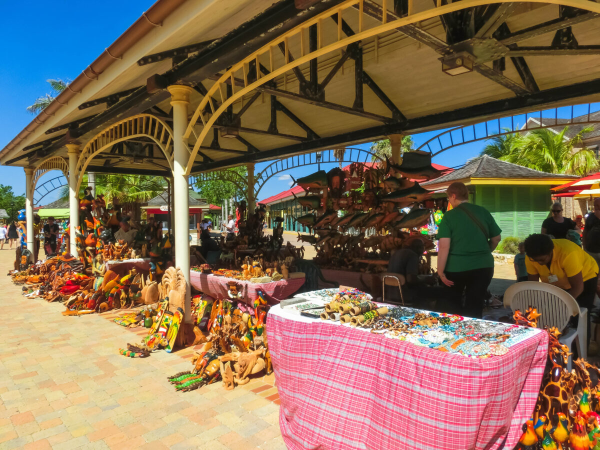 An outdoor market with stalls displaying colorful handicrafts under a roof with arched supports, in a sunny tropical setting.