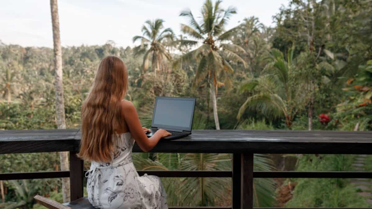 A woman with long hair works on a laptop outdoors, overlooking a tropical forest with palm trees.