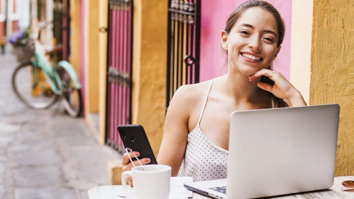 Smiling woman using a smartphone and laptop at an outdoor café table, with a bicycle in the background.