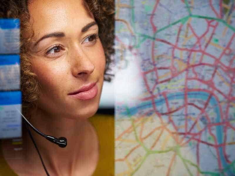 A customer service representative with a headset smiles gently, possibly ready to assist clients, with a blurred background featuring a colorful map, possibly symbolizing global connectivity and navigation.