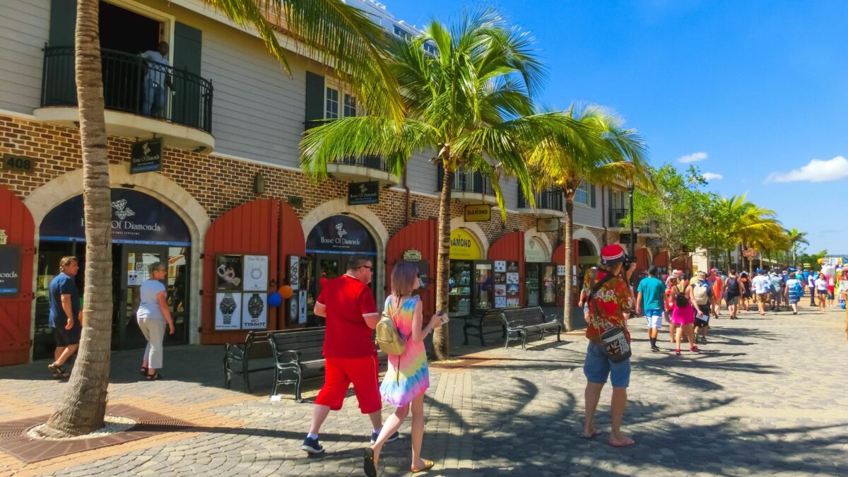 A bustling tropical street scene with tourists walking, palm trees, and shopfronts displaying jewelry, under a bright blue sky.