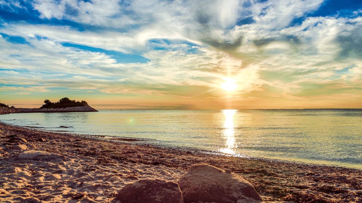 A serene sunset view at the beach with the sun reflecting on the calm water, a small island with trees on the horizon, and a sandy shore with rocks in the foreground.