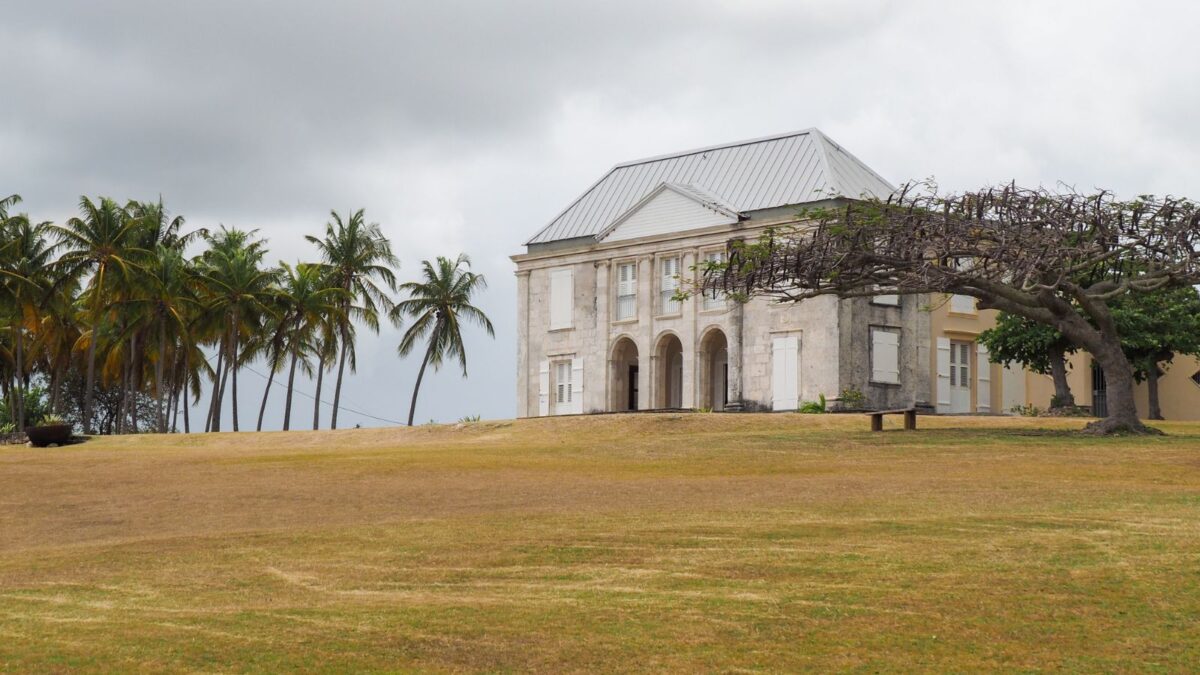 A grand stone house on a grassy hill, with palm trees and a cloudy sky in the background.