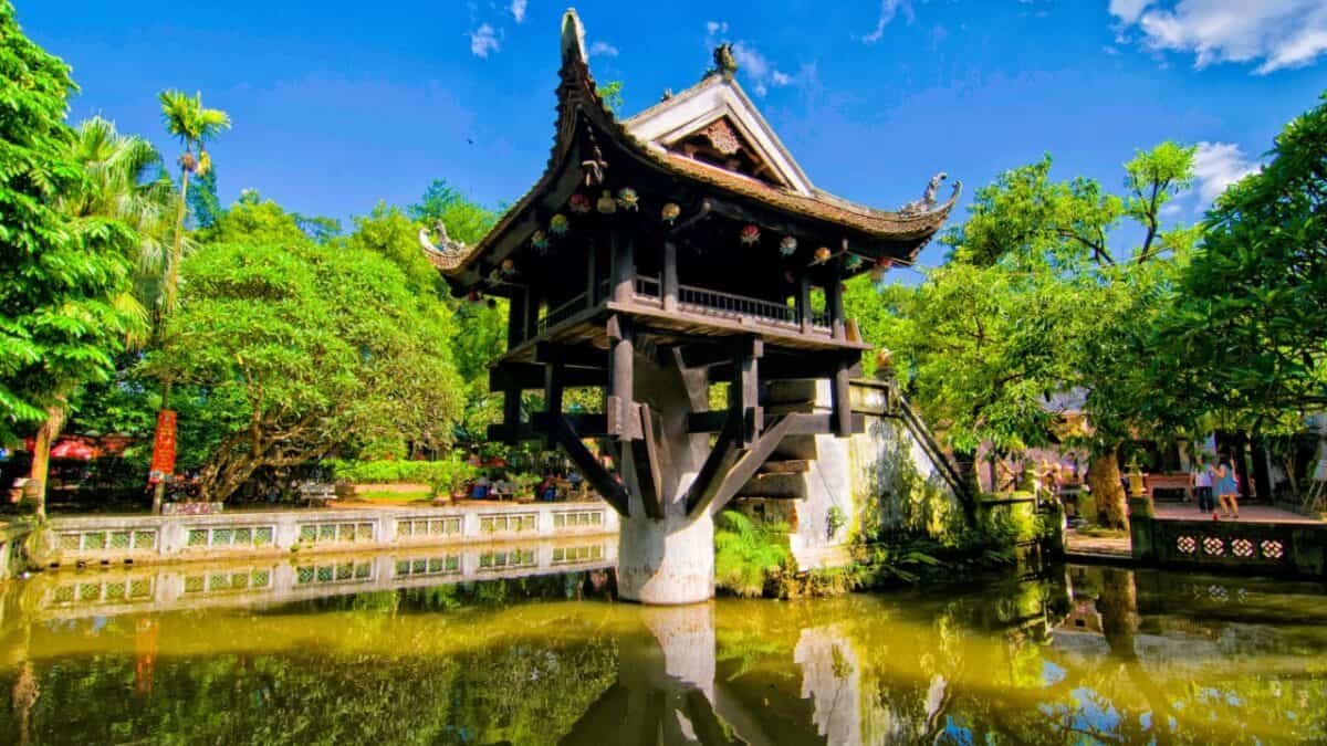 A traditional asian pagoda perched elegantly over a tranquil pond, surrounded by lush greenery under a clear blue sky.