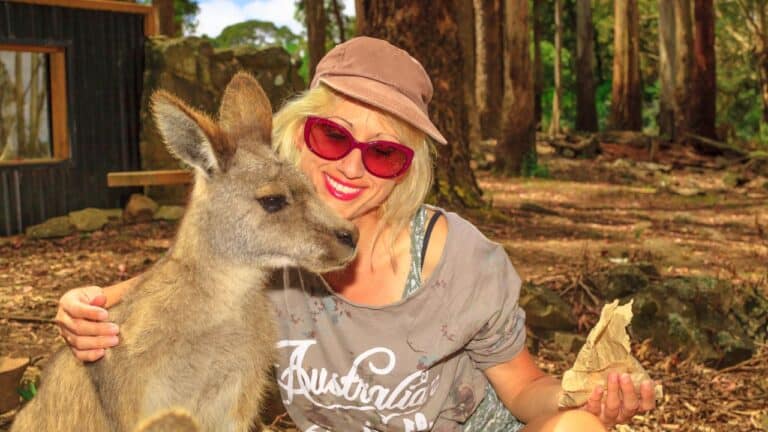A woman in a casual outfit and hat smiles while sitting next to a kangaroo in a wooded area.