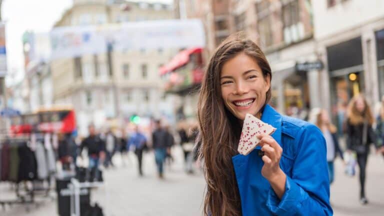 Woman in blue coat smiles while eating a sandwich on a busy city street.