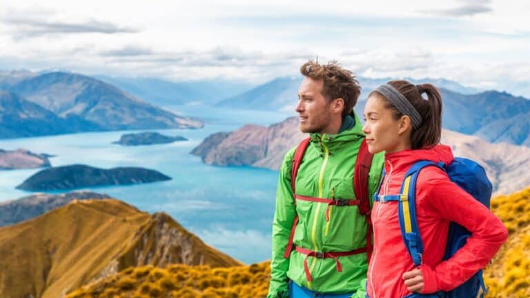 Two hikers with backpacks stand on a mountain, overlooking a scenic view of lakes and mountains under a cloudy sky.