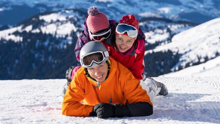 Three people in winter gear happily lying on snowy ground with mountains in the background.