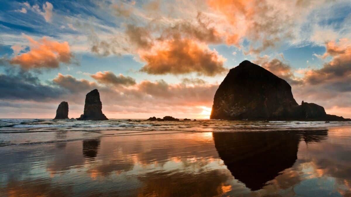 Iconic Haystack Rock at the beach during a colorful sunset with reflections on the wet sand and dramatic clouds.