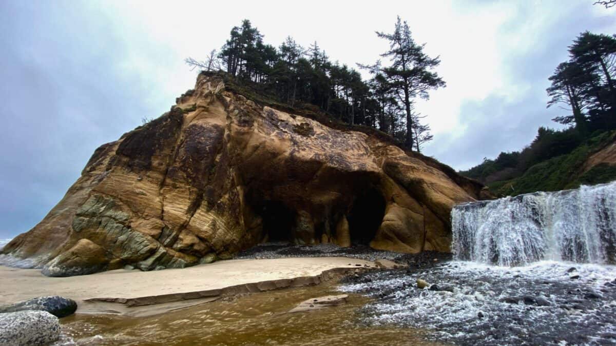 Coastal cave at Hug Point with a waterfall cascading down to a sandy beach, flanked by mossy rocks and a tree-lined cliff.