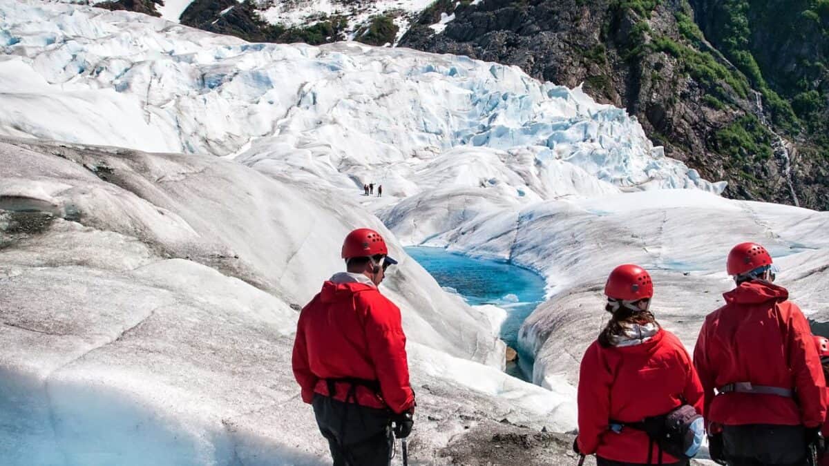 Adventurers in red jackets and helmets equipped with walking sticks exploring a rugged glacier with a visible blue ice pool.