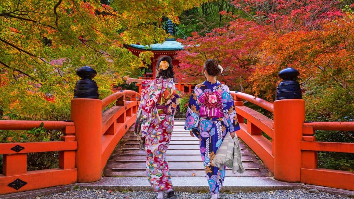 
Two women in colorful kimonos walking across a traditional red bridge in a Japanese garden during autumn.