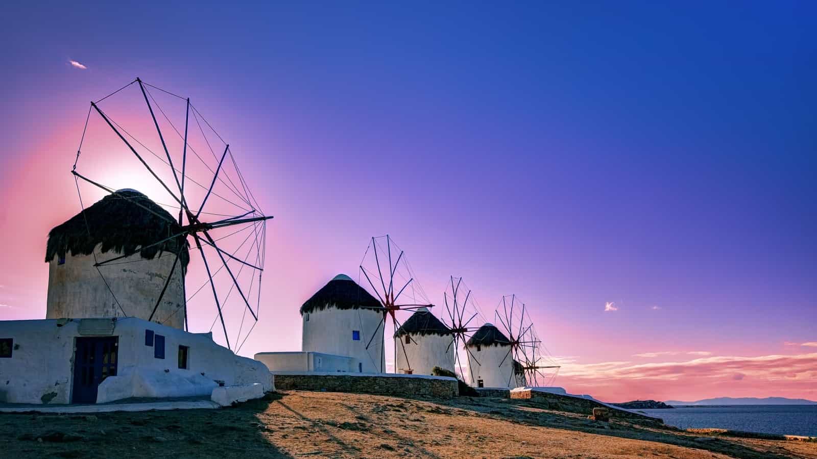 Best Greek Island for Romantic Couples 7 This is a scenic image taken during twilight, showcasing the iconic windmills of Mykonos. The sky behind the windmills is painted in soft hues of purple, pink, and blue, indicating either dawn or dusk. The windmills, with their cylindrical bases, white facades, and thatched roofs, are silhouetted against the colorful sky. Their sails are poised as if in mid-movement, adding to the dynamic of the still image. In the background, the horizon is visible along with the serene sea, contributing to the tranquil yet historic feel of the photograph.