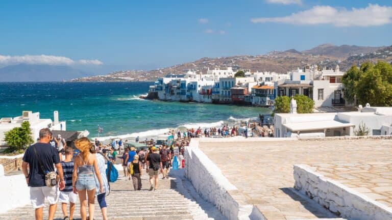 Tourists walking towards the iconic waterfront of Little Venice in Mykonos, Greece, with traditional white buildings and the Aegean Sea in the background.