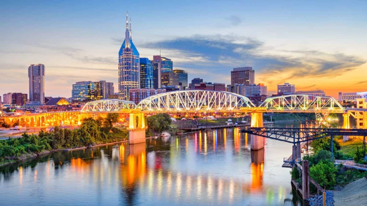 Twilight descends on a vibrant cityscape, with a distinctive skyscraper punctuating the skyline, as a steel truss bridge spans calmly over a reflective river.