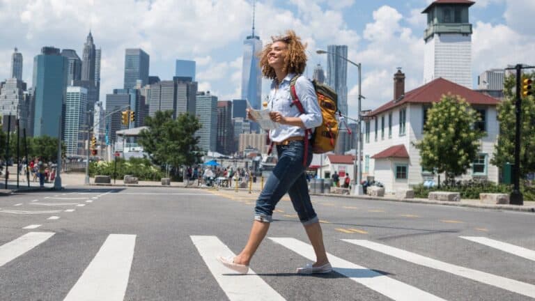 Woman with a backpack crossing a street in an urban area with skyscrapers in the background on a sunny day.
