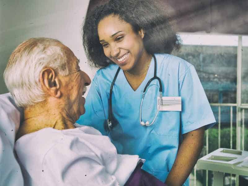 A smiling healthcare professional in scrubs and a stethoscope providing care and comfort to an elderly patient.