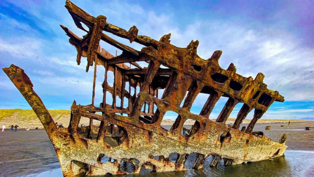 The rusted remains of the Peter Iredale shipwreck stand starkly against the sky on a sandy beach, with its skeletal structure protruding upwards, surrounded by small pools of seawater.