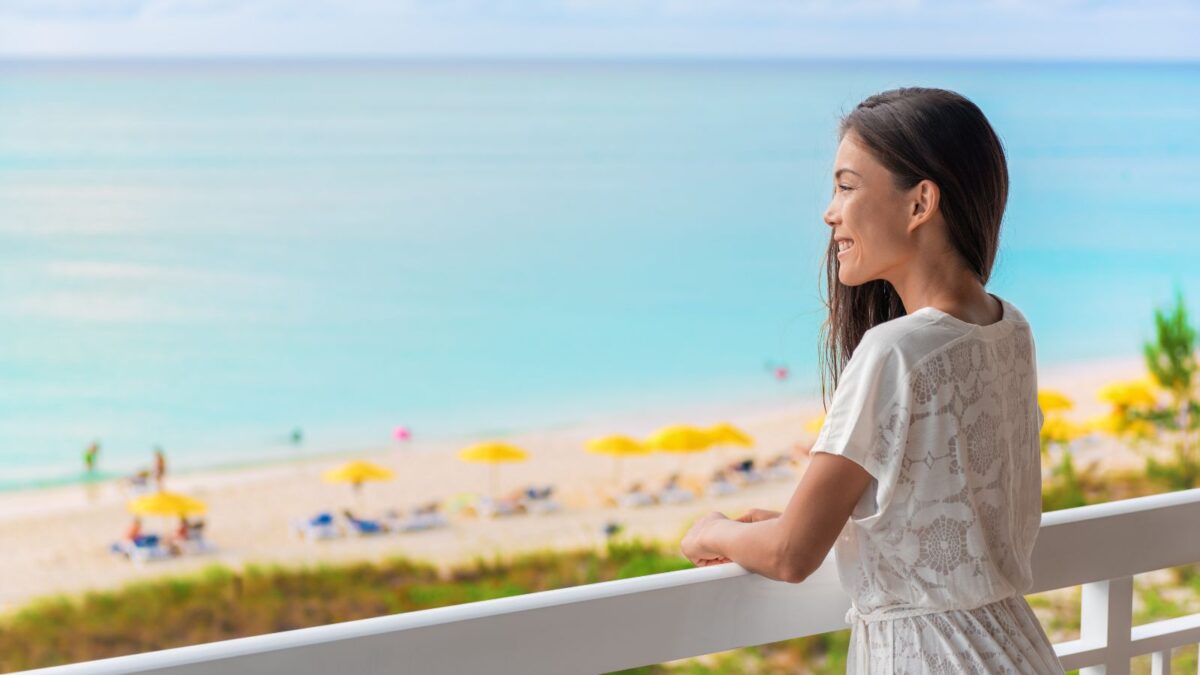 A smiling woman leaning on a balcony railing with a scenic view of a sandy beach and turquoise sea in the background.