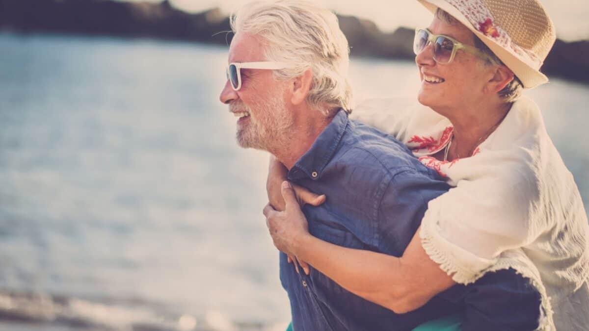 Senior couple enjoying a piggyback ride on a sunny beach day.