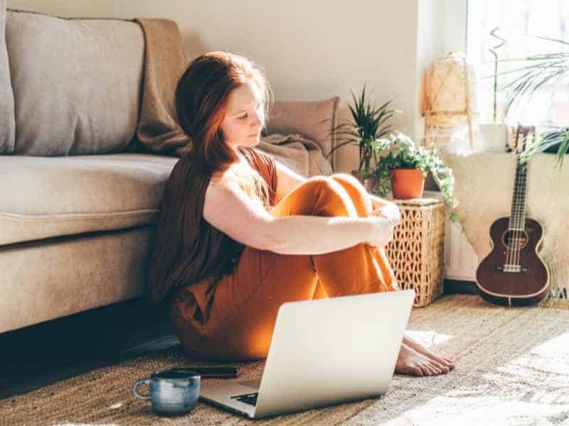 Inside the Narcissist's Playbook: 20 Tactics to Control and Confuse 12 A woman sits on the floor by her couch, working on a laptop in a sunlit room with signs of childhood neglect that resonate deeply in adulthood and a houseplant nearby.