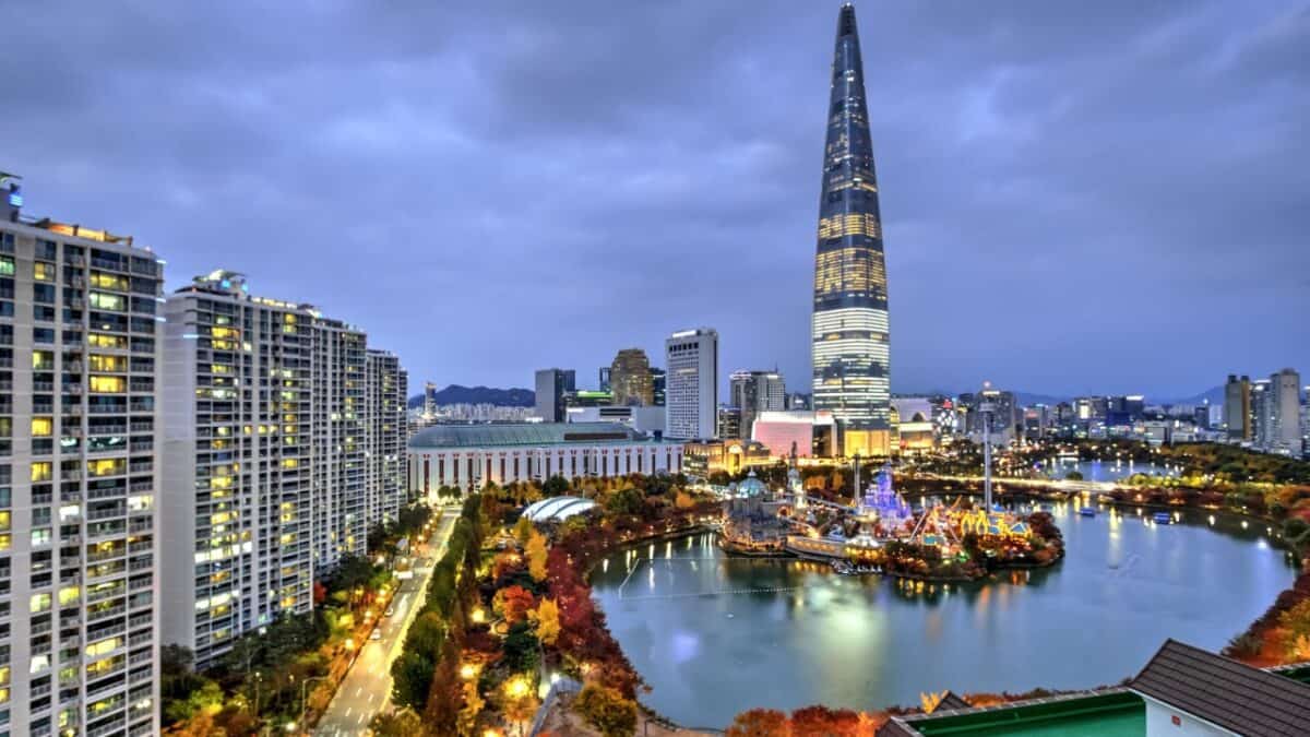 20 Best Places For Solo Female Travelers 23 A cityscape at dusk featuring a prominent, futuristic skyscraper towering above the surrounding buildings, with a serene park and lake illuminated by warm lights in the foreground and a majestic mountain backdrop under a cloudy sky.