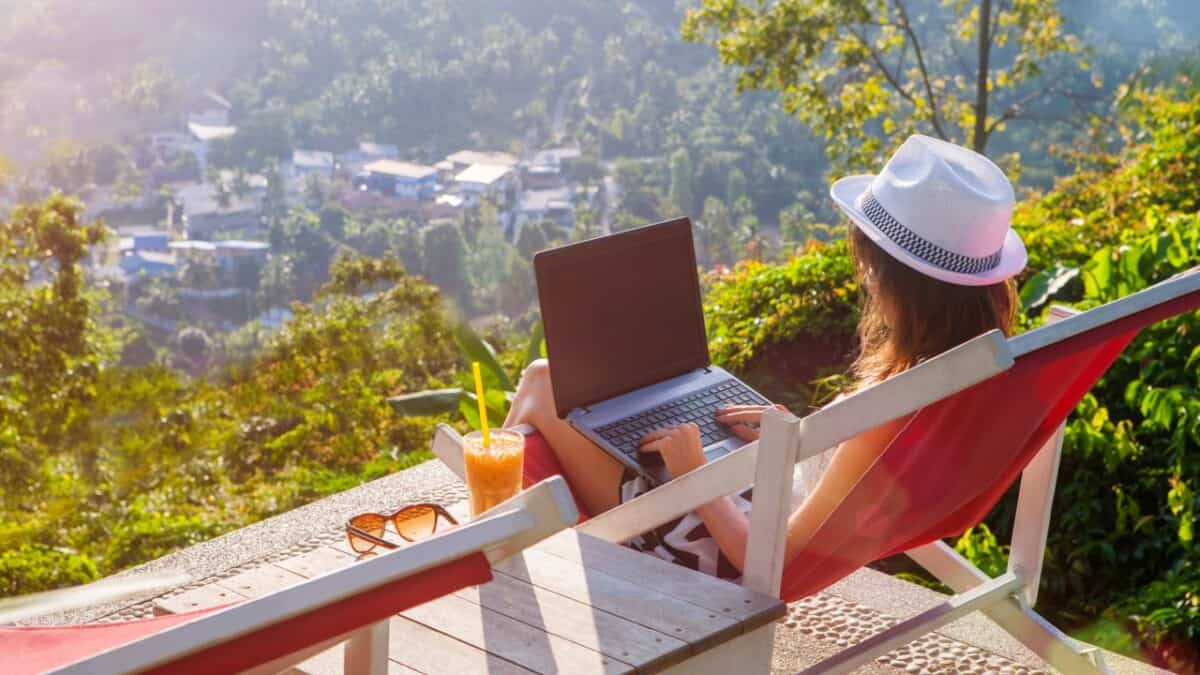 Woman in a sunhat on a red deck chair using a laptop overlooking a scenic village with a drink and sunglasses beside her.