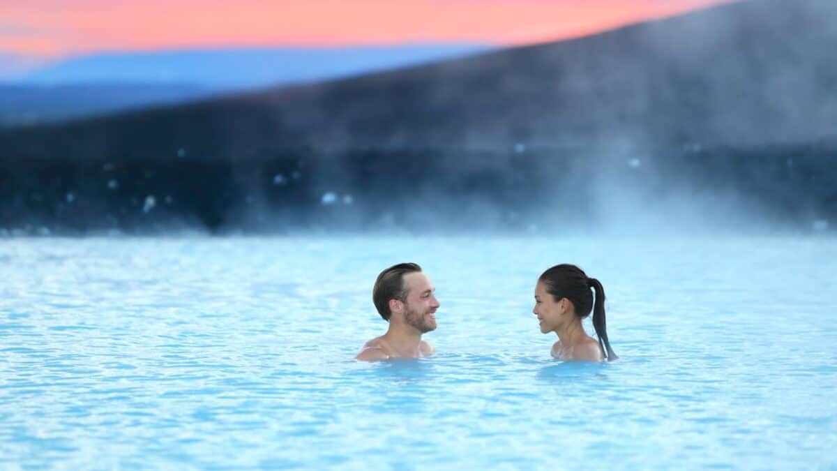 A man and a woman enjoying a warm, steaming geothermal spa, the Blue Lagoon, with a sunset creating pink hues in the sky in the background.