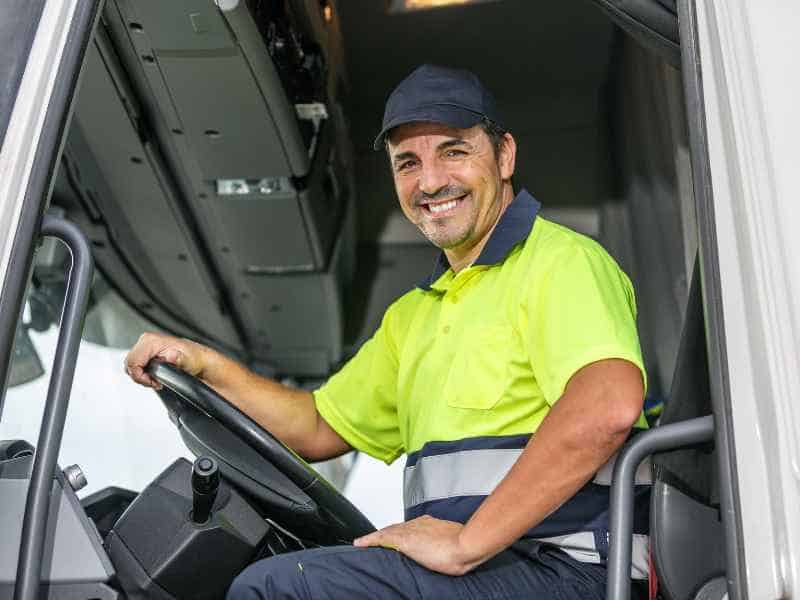 A cheerful truck driver in a high-visibility shirt and cap sitting behind the wheel of his vehicle, ready to start his route.