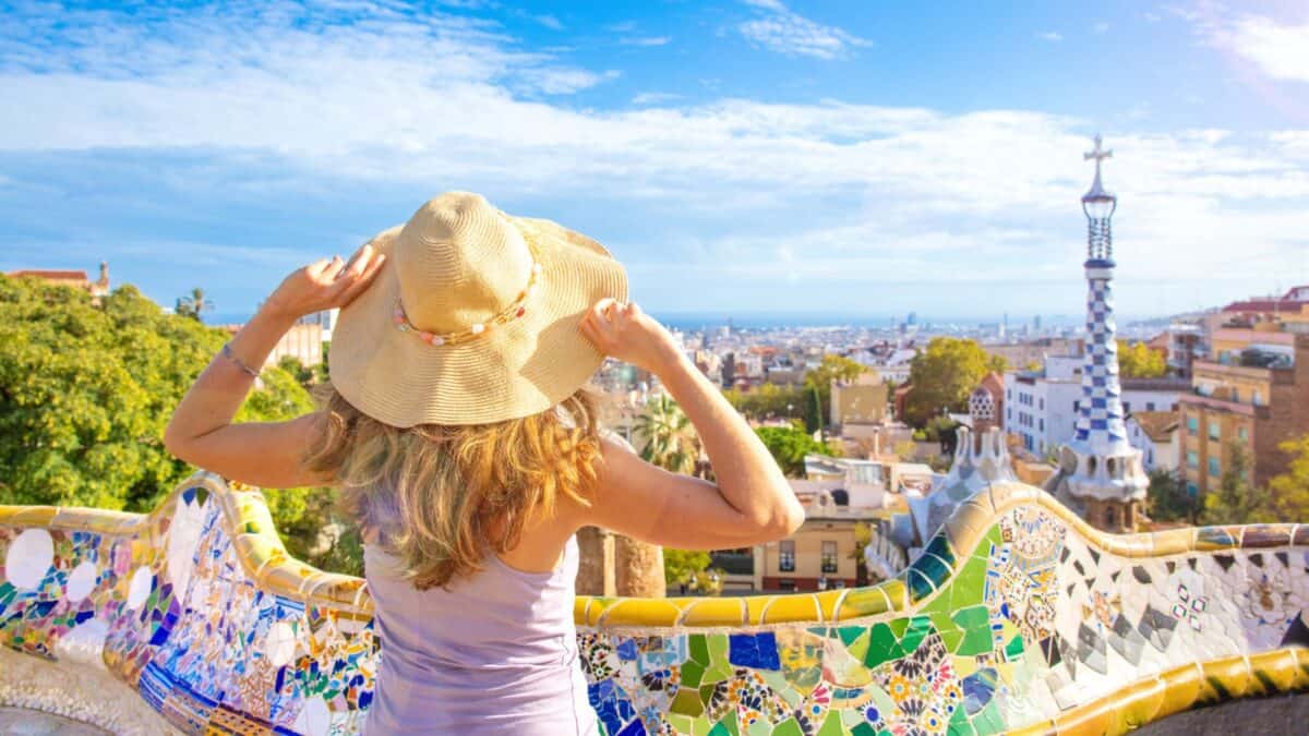 20 Best Places For Solo Female Travelers 16 A woman wearing a straw hat views the colorful mosaic benches and vibrant skyline of barcelona from park güell on a sunny day.