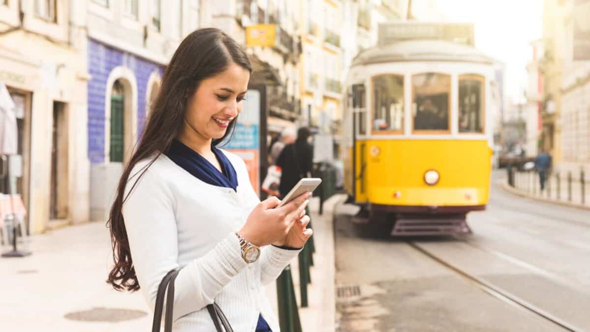 20 Best Places For Solo Female Travelers 9 A young woman smiling while looking at her smartphone, standing on a city street with a yellow tram passing by in the background.