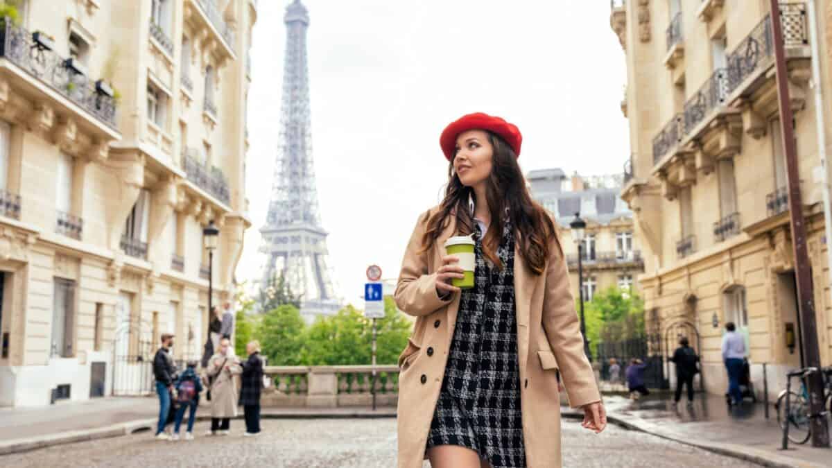 15 Top Travel Destinations for Moving On After a Breakup 3 A stylish woman wearing a red beret and trench coat walks through a parisian street with a coffee cup, with the eiffel tower in the background.