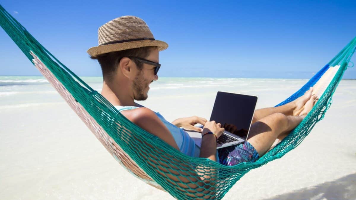 A man wearing sunglasses and a hat is using a laptop while relaxing in a hammock on a beach.