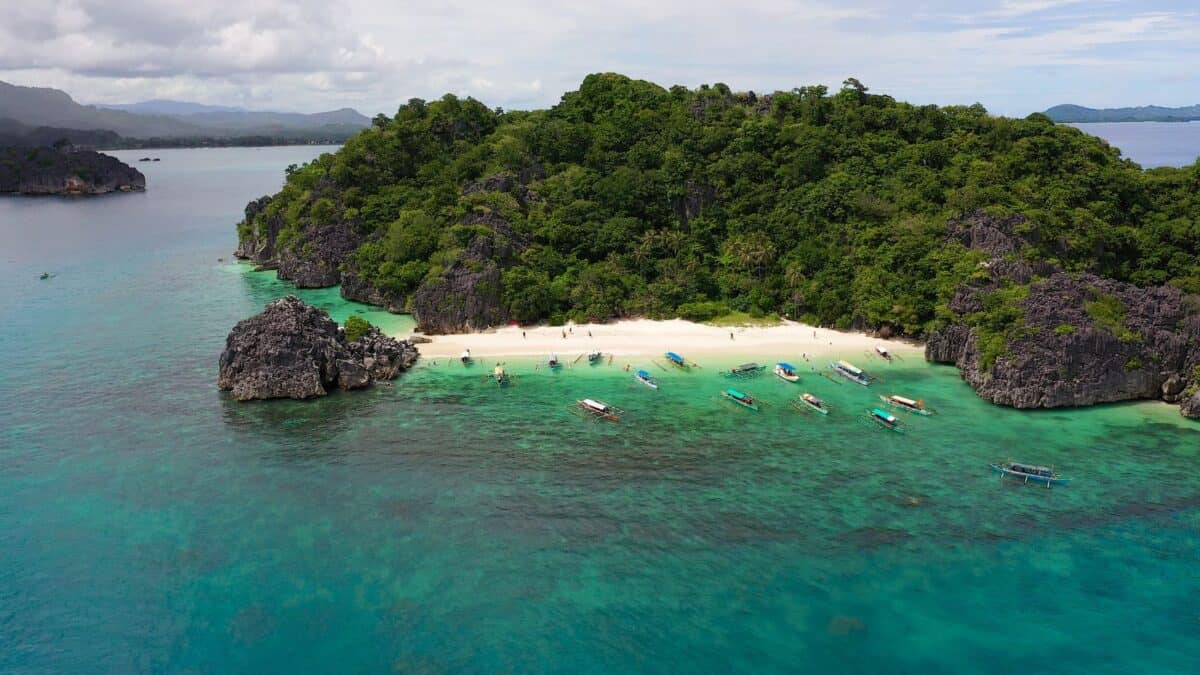 An aerial view of a tropical beach with boats, surrounded by lush greenery and clear turquoise waters.