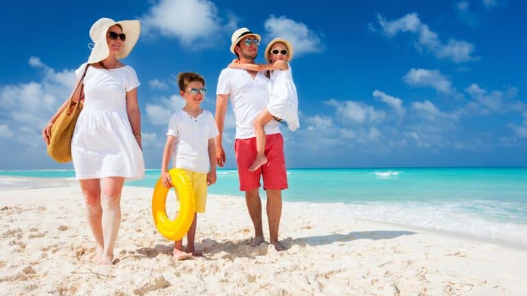 A family of four, with a mother, father carrying a young child, and a boy holding a yellow float, all dressed in summer clothes, walking on a white sandy beach with the clear blue sea in the background.