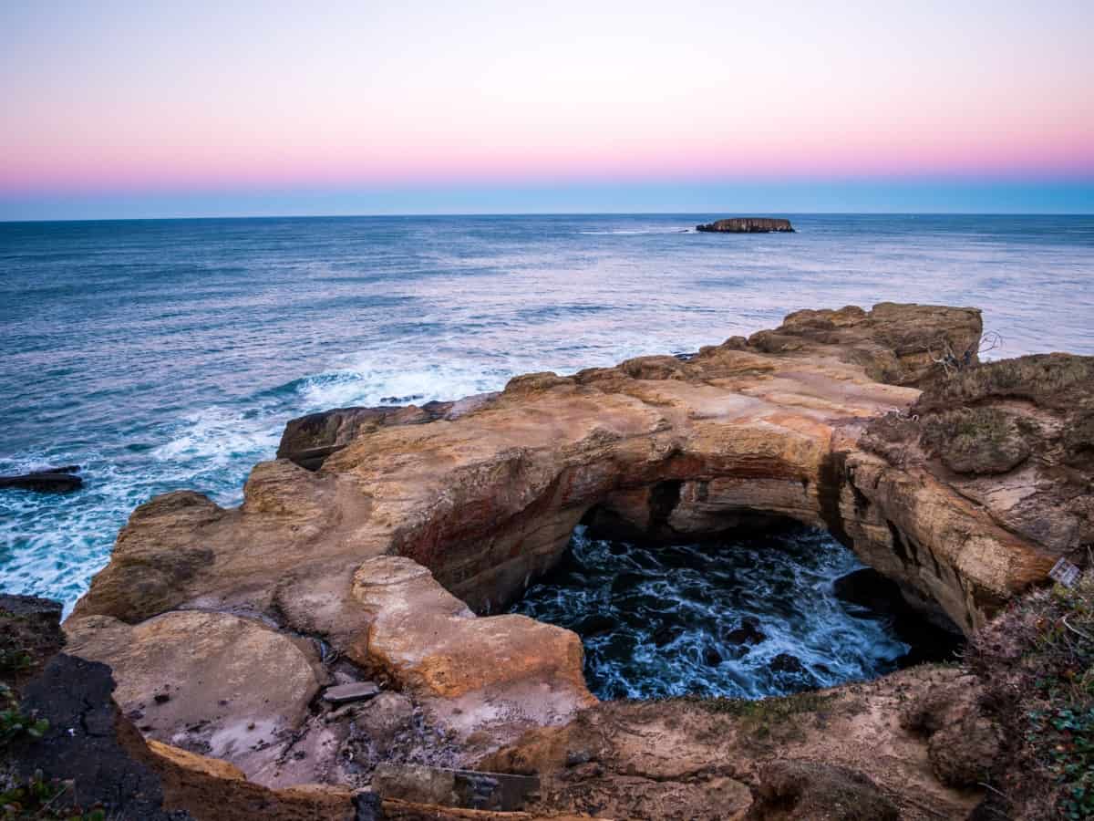 The geological wonder of Devil's Punchbowl on the Oregon coast, captured at twilight; the natural sandstone arch encircles a turbulent sea, under a sky softly blending pink and blue hues.