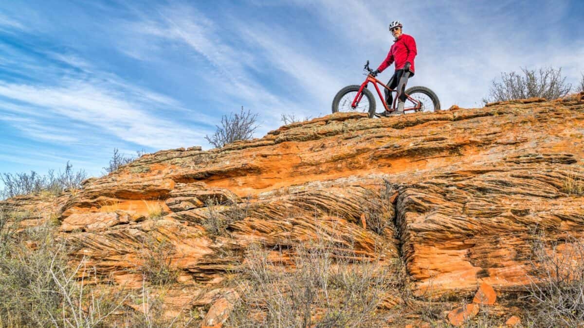 Cyclist with a fat bike standing on striated orange rock formations under a blue sky with wispy clouds.