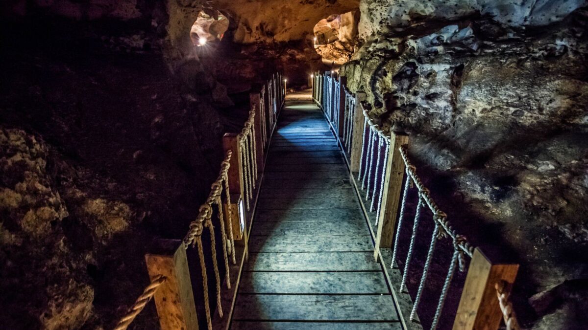 A wooden walkway with rope handrails leading through a dimly lit, rugged cave.