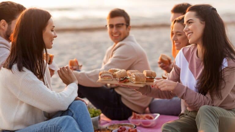 A group of friends enjoying a picnic with sandwiches on the beach at sunset.