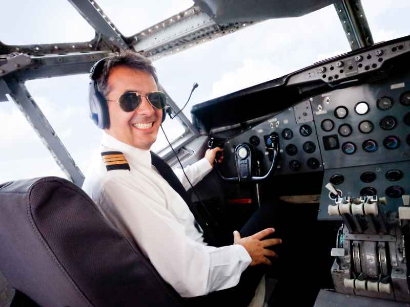Pilot in uniform smiling confidently at the camera while sitting in the cockpit of an airplane, with his hand on the controls.