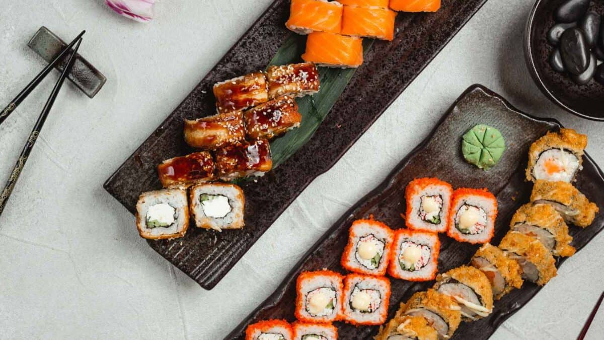 Assorted sushi and rolls plated on antique black rectangular dishes, accompanied by soy sauce and chopsticks on a textured gray surface.